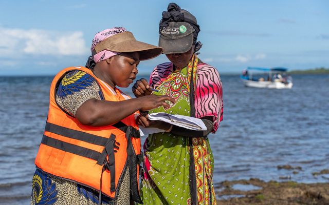 A woman, right, wearing a TNC baseball hat, stands on a beach and holds a notebook. Another woman, left, is pointing at something in the notebook.