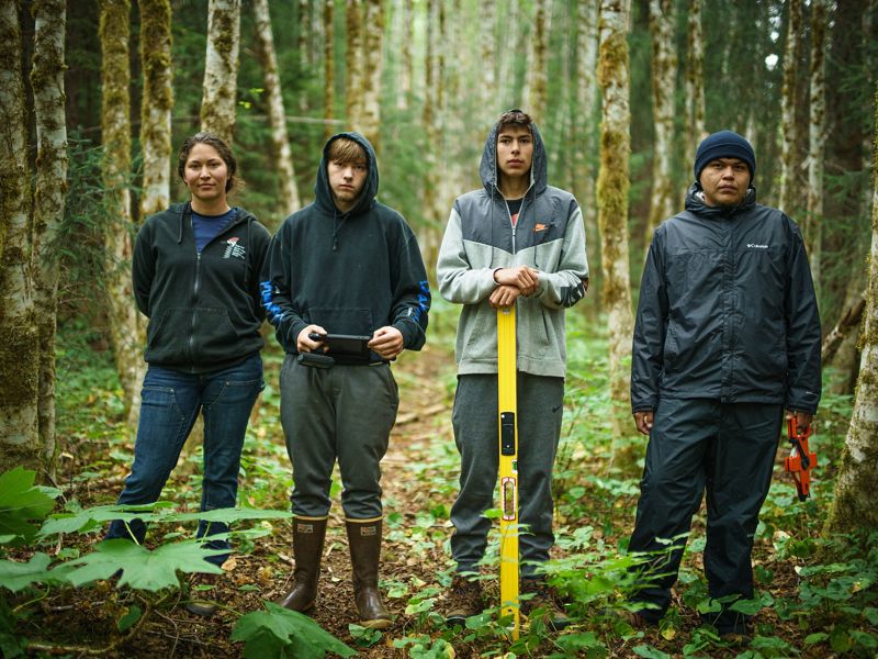 4 young adults wearing boots and jackets stand in a forest while holding various tools.