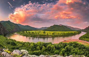 A wide river curves between lush green banks. Mountain peaks rise in the background under clouds that glow golden in the setting sun.