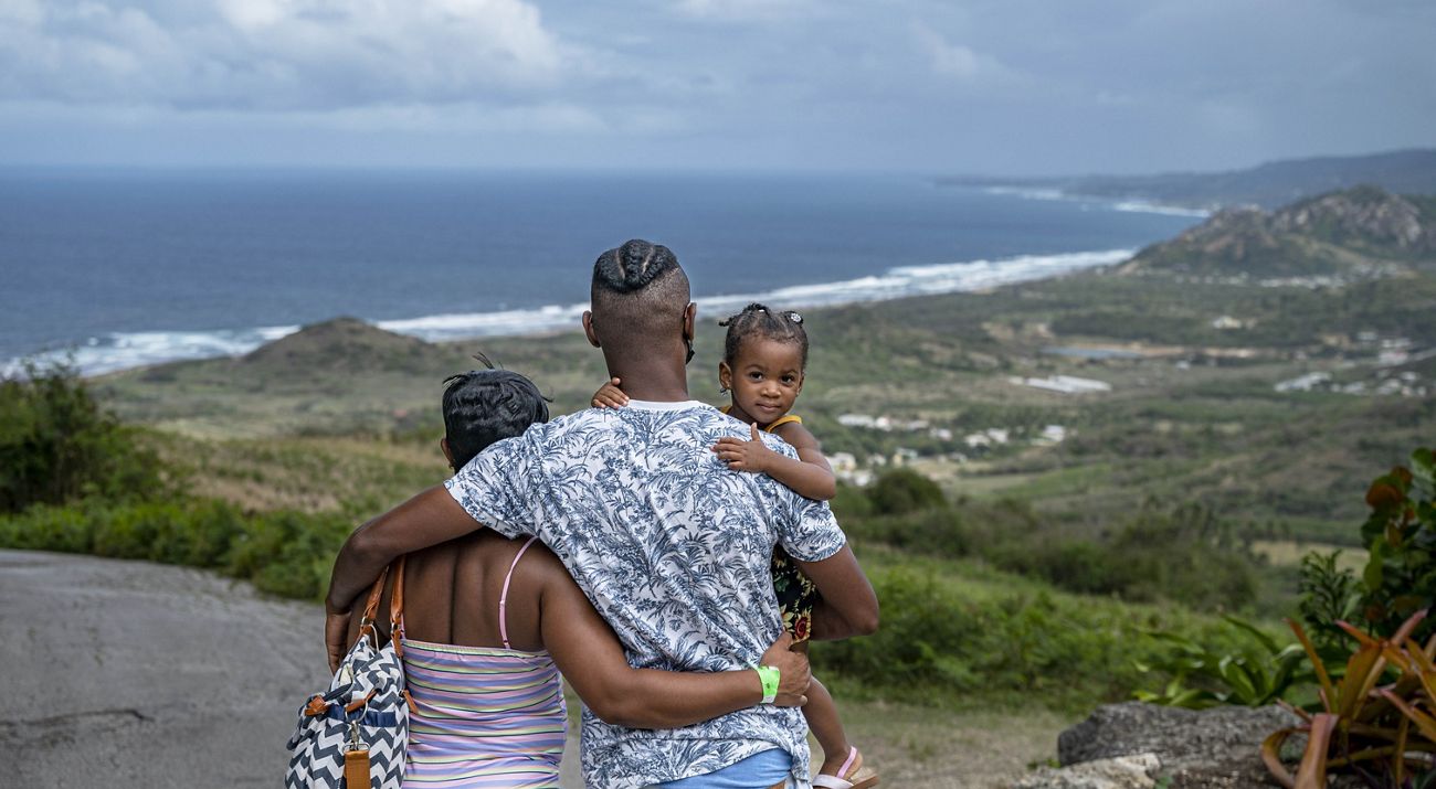 Photo of a family - father and mother holding young child - looking out over a Barbados hilltop toward the ocean.