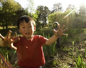 smiling toddler stands in a green space with mud on his hands
