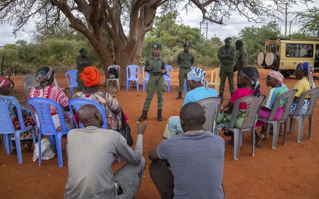 A group of people sit in a circle to speak with several park rangers, standing in the middle of the circle.