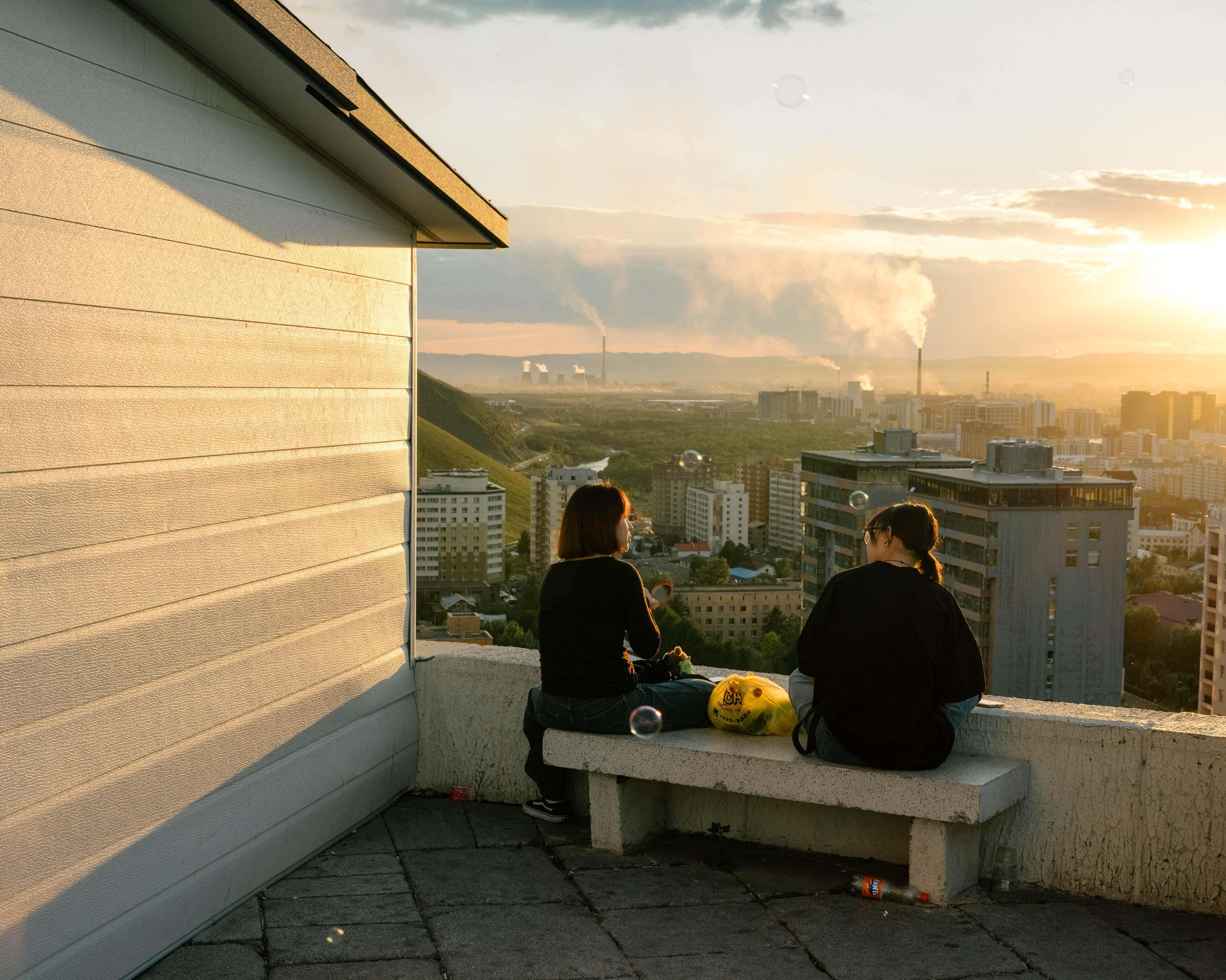 A couple of women sitting on a bench overlooking a city.