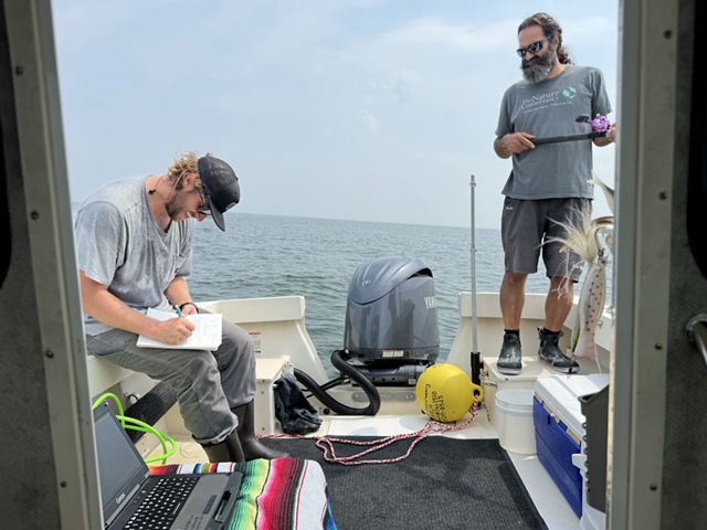 Two men working on a boat. One is standing on the right one is sitting on the left writing in a notebook. 