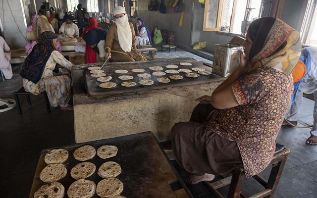 A woman tends to dozens of flat breads on a large stove as other community members work in the background.