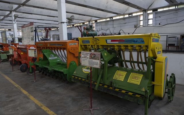 In a long university building, three large seeder machines of different colors sit next to each other, full of rotors and hoses.