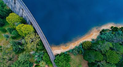 Aerial view of a large, vibrant-blue body of water behind a narrow dam wall, with forests on the banks of the water and on the other side of the dam.